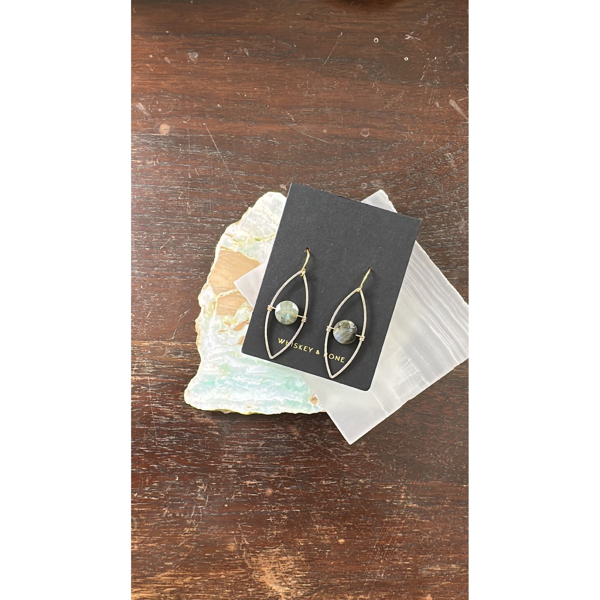 Pair of gold earrings on a black card with 'Whiskey & Bone' branding, placed on a wooden surface.
