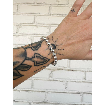 Hand with silver beaded bracelet and tattoo on wrist against a white brick wall background