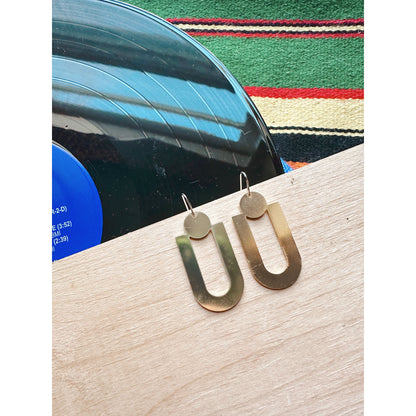 Gold dangle earrings on a wooden surface with a colorful cloth in the background