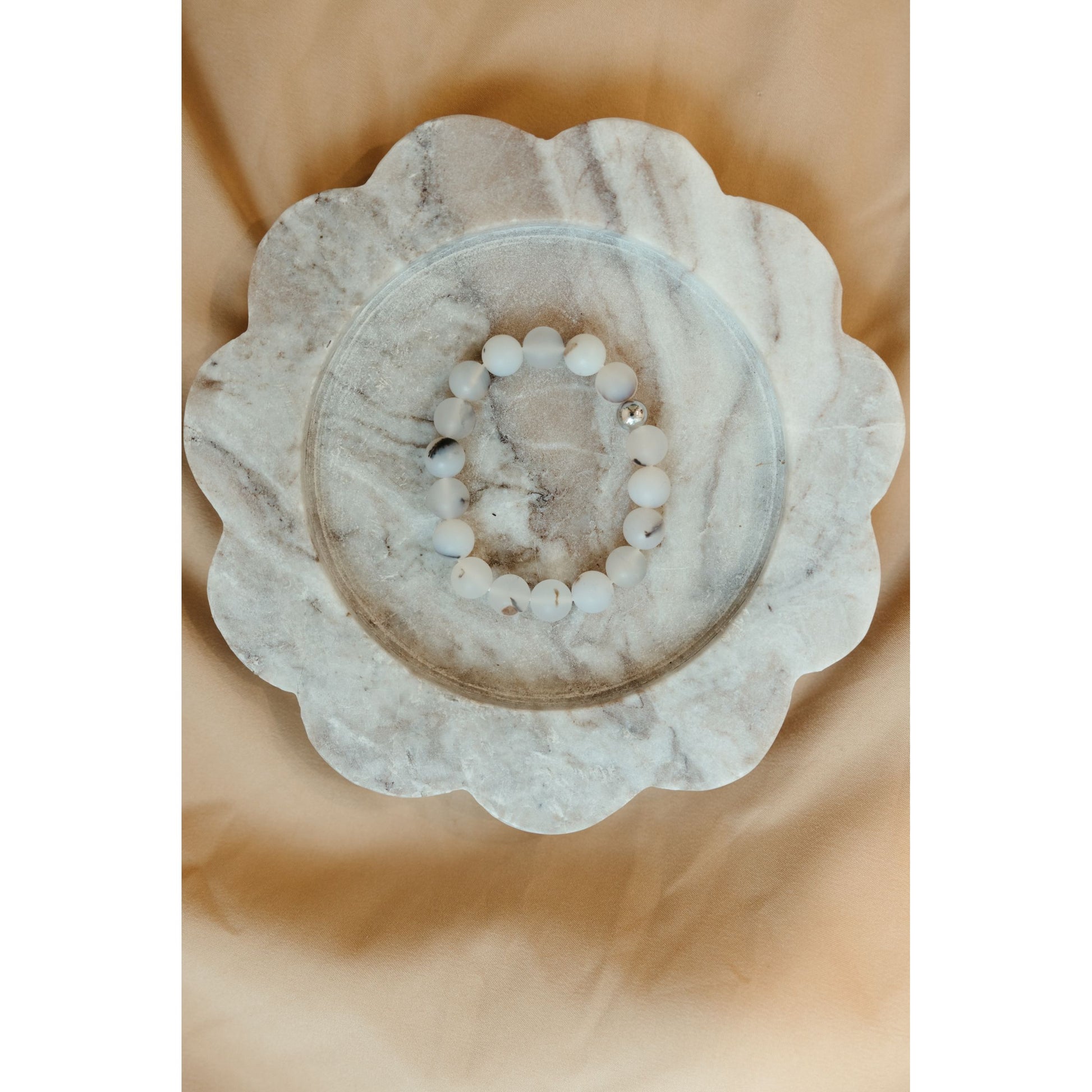 A beaded bracelet made of white and light gray round beads with a few dark accents, displayed on a scalloped white marble tray over a soft beige fabric background.
