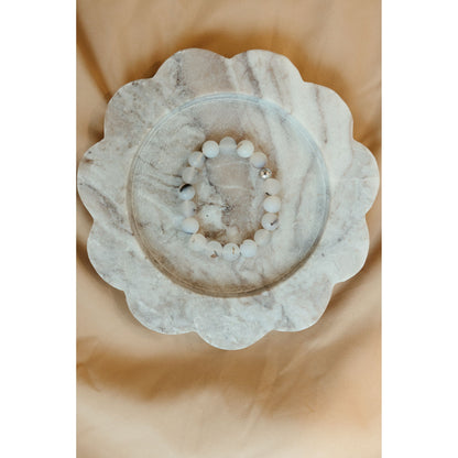 A beaded bracelet made of white and light gray round beads with a few dark accents, displayed on a scalloped white marble tray over a soft beige fabric background.