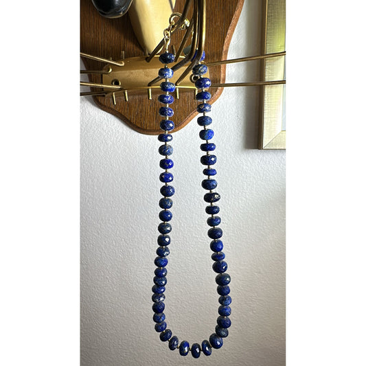 Blue beaded necklace hanging on a wooden stand against a white wall.