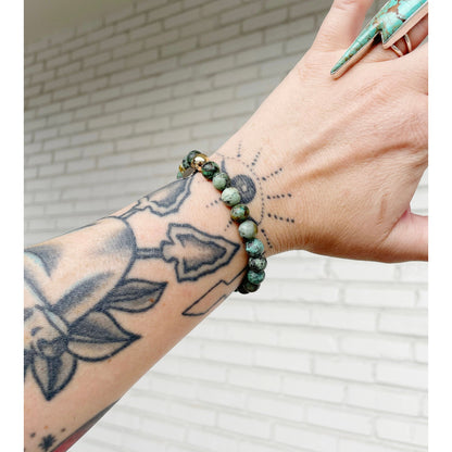 Person wearing a green beaded bracelet on a tattooed wrist with a white brick wall background